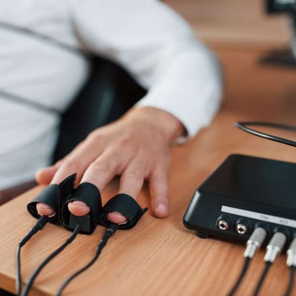 Hand with electrodes on fingers is on the table. Suspicious man passes lie detector in the office. Asking questions. Polygraph test.
